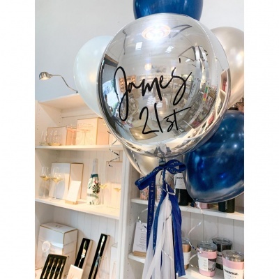 Balões em prata metálico, azul, branco e bege com texto 'James 21st' e fitas penduradas em loja