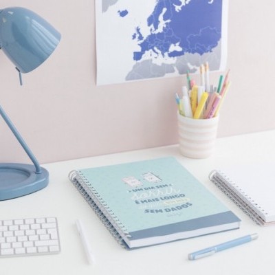 Mesa de escritório com cadernos, candeeiro azul, teclado branco e material de escrita colorido.