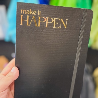 Caderno preto com texto dourado 'make it HAPPEN' e elástico na capa.