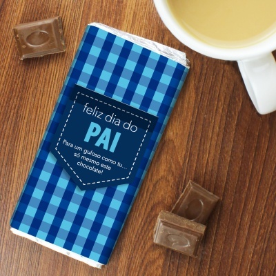 Barra de chocolate embrulhada com padrão azul e texto comemorativo sobre mesa de madeira com pedaços de chocolate e chávena.