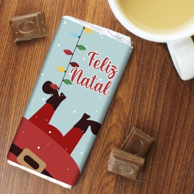 Barra de chocolate embrulhada com texto 'Feliz Natal' e decoração de Natal sobre mesa de madeira, com pedaços de chocolate e chávena próxima.