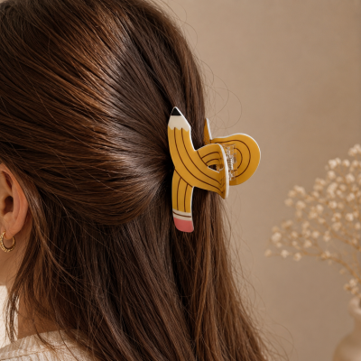 Prendedor de cabelo amarelo com design de lápis, preso em cabelo castanho.