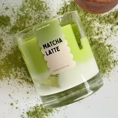 Copo de vidro com bebida branca e verde etiquetada como Matcha Latte sobre fundo com pó verde.