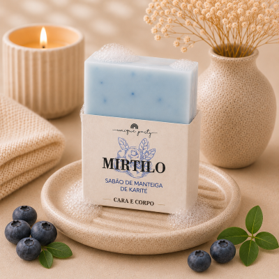 Sabonete azul claro com embalagem em papel creme num prato creme, ao lado de mirtilos, toalha, vela acesa e vaso com flores secas