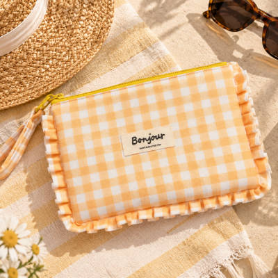 Bolsa xadrez amarela e branca com etiqueta Bonjour sobre toalha amarela, chapéu de palha, óculos escuros, limão e copo de bebida com limão