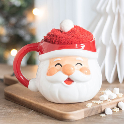 Caneca de cerâmica com formato da cara do Pai Natal, com chapéu vermelho com pompom branco, em cima de tábua de madeira com mini marshmallows e estrelas de açúcar.