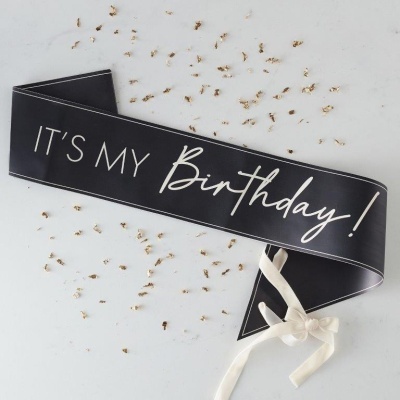 Faixa preta com texto branco 'IT'S MY Birthday!' com confetes dourados e fita branca