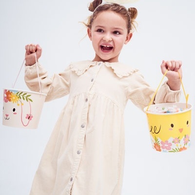 Criança com vestido creme e tiara de coelho segurando baldes decorados coloridos.