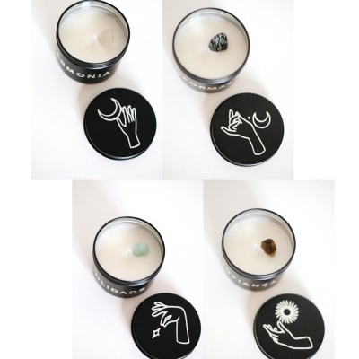 Quatro velas brancas em recipientes de vidro preto com tampas ilustradas e pedras no centro