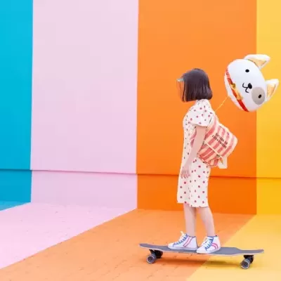 Menina em vestido com bolinhas vermelhas a andar de skate com mochila e balão de cão coloridos