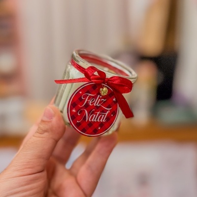 Vela branca em frasco de vidro com etiqueta 'Feliz Natal' e laço vermelho
