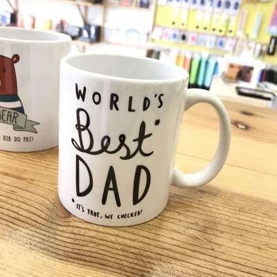 Caneca branca com texto 'WORLD'S Best DAD' em preto sobre mesa de madeira