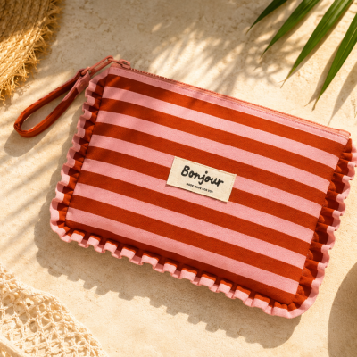 Necessaire às riscas vermelha e rosa com franja, ao lado de chapéu de palha, óculos escuros, folhas verdes e melancia.