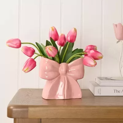 Vaso rosa em forma de laço com tulipas cor-de-rosa sobre mesa de madeira clara