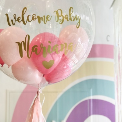 Balão transparente com interior de balões cor-de-rosa e texto dourado de boas-vindas para bebé