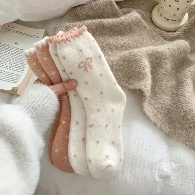 Três pares de meias tricotadas em rosa e branco com padrões de bolinhas e laço, seguradas sobre roupa e cama brancas e castanhas.