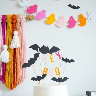Bolo branco decorado com morcegos pretos e letras coloridas, decoração de Halloween na parede.