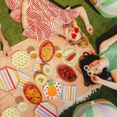 Piquenique colorido com toalha, pratos e guardanapos, snacks e três pessoas ao sol
