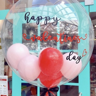Balão transparente com balões interiores rosa e vermelho e texto 'happy valentines day' na frente de porta de vidro azul