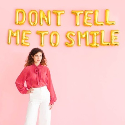 Mulher com blusa rosa e calças brancas diante de parede rosa com letras douradas de balão