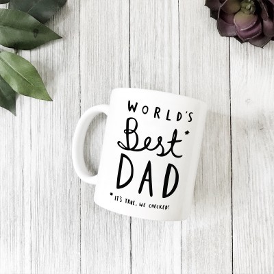 Caneca branca com texto 'WORLD'S Best DAD' num fundo de madeira clara com folhas verdes e planta suculenta.