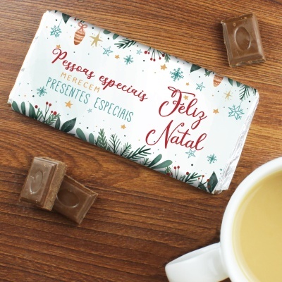 Barra de chocolate embrulhada com tema de Natal sobre mesa de madeira ao lado de pedaços de chocolate e chávena