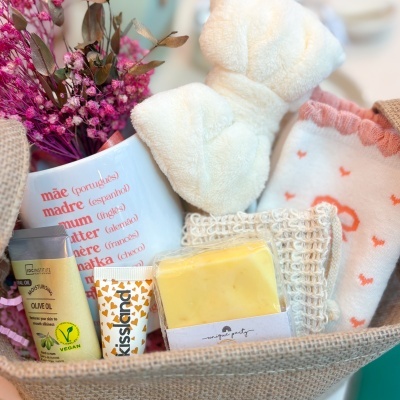 Conjunto de presente com meias, faixa de cabelo, sabonete, óleo vegano, fita esfoliante e copo com texto em várias línguas, em caixa de tecido bege.