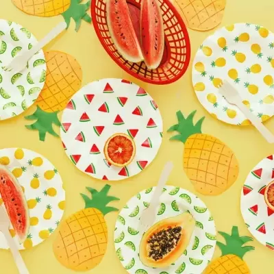 Pratos descartáveis com padrões de frutas e cestos vermelhos com snacks sobre fundo amarelo decorado com recortes de abacaxi.