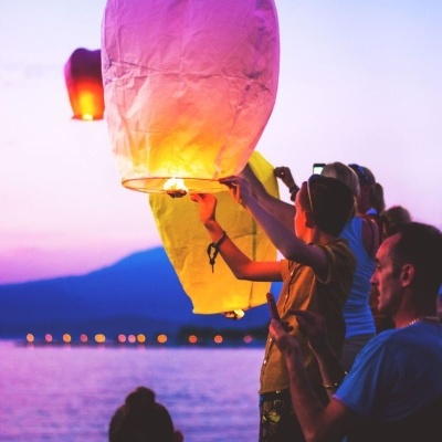 Pessoas lançando lanternas celestes de papel coloridas sobre a água ao entardecer