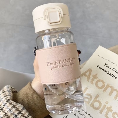 Garrafa de água transparente com capa rosa clara e texto HanXPARSON, segurada na mão, com livro ao fundo.