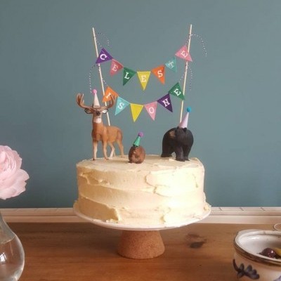 Bolo branco com decoração de animais e faixa colorida em suporte de cortiça numa mesa de madeira