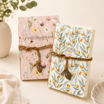 Dois cadernos decorativos com padrões de flores e citrinos, fechados com cordão castanho e pendente de folha.