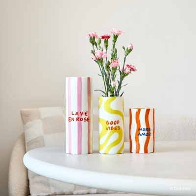 Três vasos cerâmicos com padrões coloridos e textos em inglês e francês sobre uma mesa branca, com flores rosas num dos vasos.