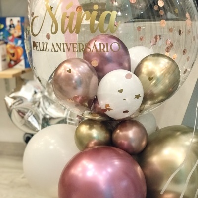 Balão transparente com texto dourado e balões metálicos rosa, dourados e brancos, em exposição numa loja
