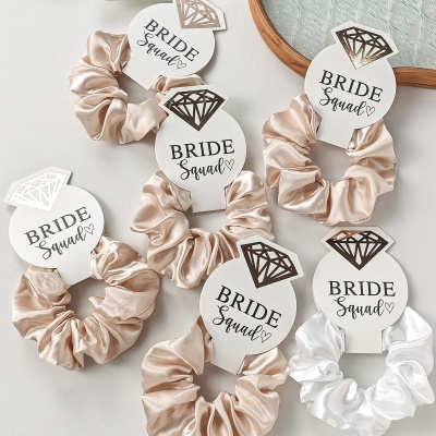 Elásticos de cabelo satinados com cartão branco e texto Bride Squad dispostos sobre superfície branca.