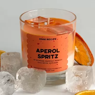 Vela em copo de vidro com cera laranja e branca, rótulo laranja APÉROL SPRITZ com cubos de gelo e fatias de laranja seca