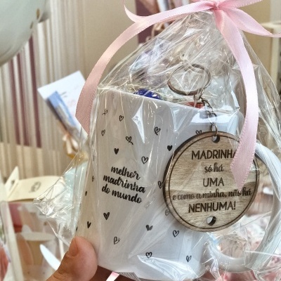 Caneca branca com estampa de corações e texto para madrinha, embrulhada em celofane com fita rosa