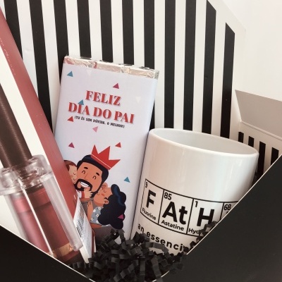 Conjunto de presente com baton rosa, barra de chocolate 'FELIZ DIA DO PAI' e caneca branca com design de tabela periódica numa caixa preta e branca.