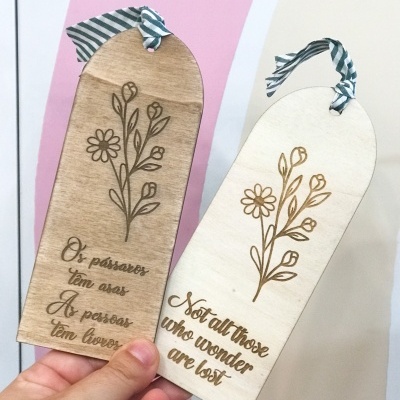 Dois marcadores de livros em madeira com ilustrações de flores e frases gravadas em português e inglês.