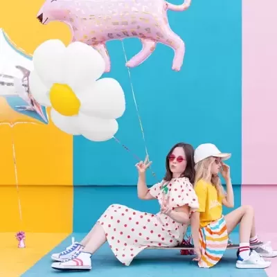 Duas raparigas sentadas com balões coloridos contra fundo em amarelo, azul e rosa.