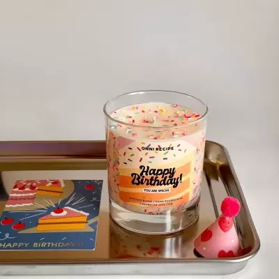 Vela de aniversário em copo de vidro com confetis coloridos e rótulo, numa bandeja metálica com cartão e chapéu de festa.