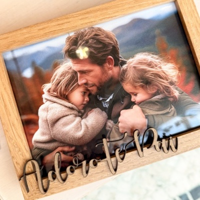 Moldura de madeira clara com fotografia de pai abraçando duas crianças e frase 'Adoro-te Pai' em madeira