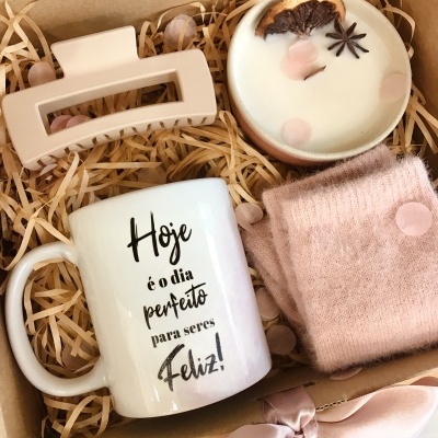 Caixa de presente com caneca branca com mensagem, vela decorativa, presilha de cabelo bege e cachecol rosa com bolinhas, em enchimento de palha artificial.
