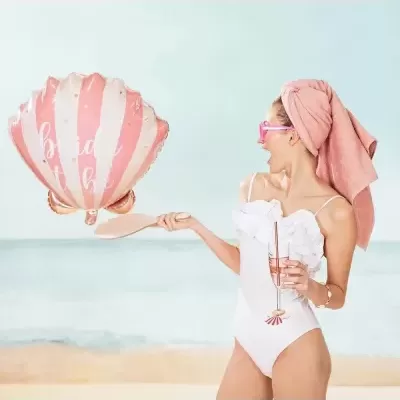 Mulher com fato de banho branco e toalha cor-de-rosa na cabeça segurando balão 'bride to be' na praia