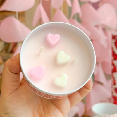 Vela branca com corações em cera cor-de-rosa e creme num recipiente redondo, segurada por mão