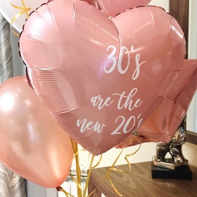 Balões cor de rosa com texto '30's are the new 20's' num ambiente interior com mesa e cortinas.