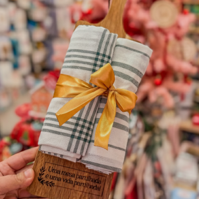 Panos de cozinha brancos com riscas verdes amarrados a uma tábua de madeira com texto gravado, em loja
