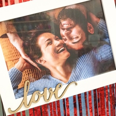 Moldura branca com fotografia de casal e palavra love em madeira sobre fundo vermelho e branco ás riscas.