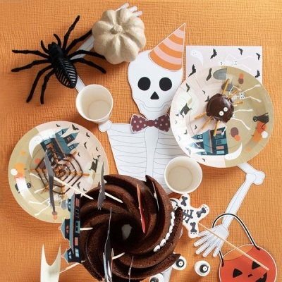 Mesa de Halloween com pratos temáticos, aranhas decorativas, abóbora branca e bolo de chocolate sobre toalha laranja.