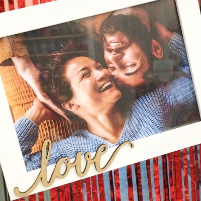 Fotografia de casal sorridente em moldura branca com palavra 'love' em madeira